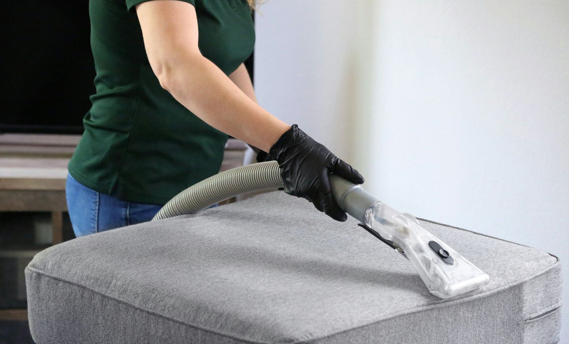 a woman in a green shirt and black gloves vacuuming a gray ottoman